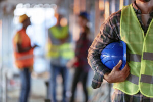 Man holding blue helmet close up. Construction man worker with office and people in background. Close up of a construction worker's hand holding working helmet.